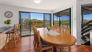 A dining area with a wooden table and chairs, a clock on the wall, and a view of a garden through a glass door. - Ideal Beach Family Getaway with Ocean Views (Noosa North Shore)