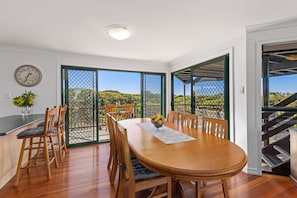 A dining area with a wooden table and chairs, a clock on the wall, and a view of a garden through a glass door.