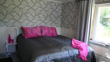 Un dormitorio con una colcha acolchada oscura, dos almohadas rosas con un estampado de árboles y una ventana con persianas.