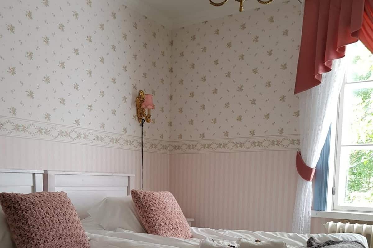 A neatly made bed with pink pillows, a white headboard, and a window with red curtains.