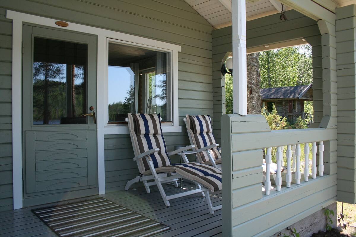 A porch with two striped chairs and a small table, a wooden door, and a view of a cabin in the background.