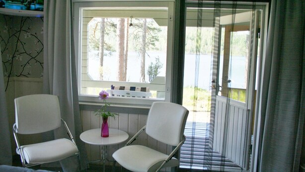 A room with two white chairs, a small round table with a vase, and a window with a view of trees. - Taulun Kartano (Toivakka)