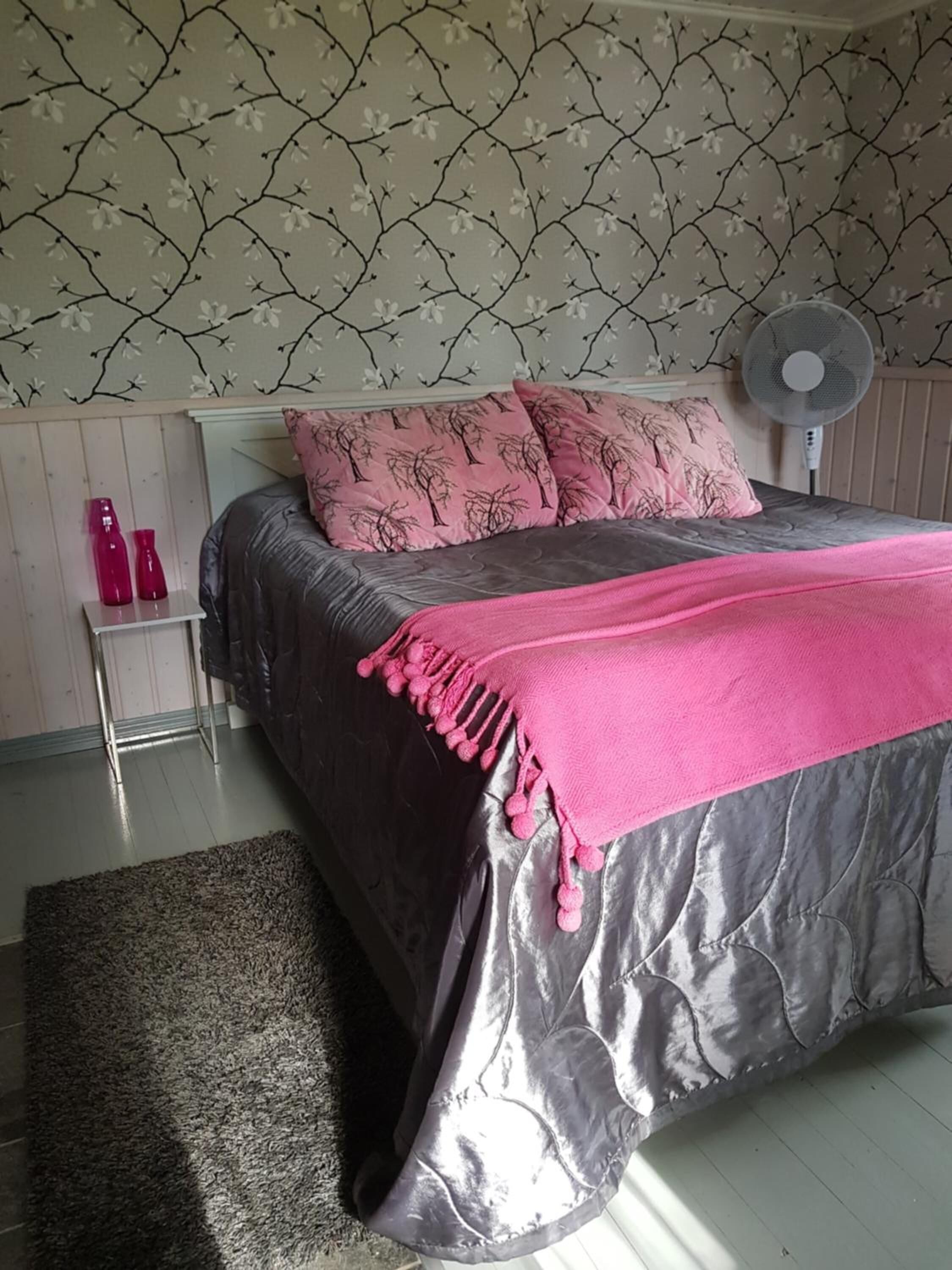 Une chambre à coucher avec un lit, une table de chevet sur laquelle sont posées deux bouteilles roses, un ventilateur mural et un papier peint à motifs.