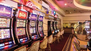 A row of slot machines in a casino with a carpeted floor and chairs in the background. - The Carousel Hotel (Hammanskraal)