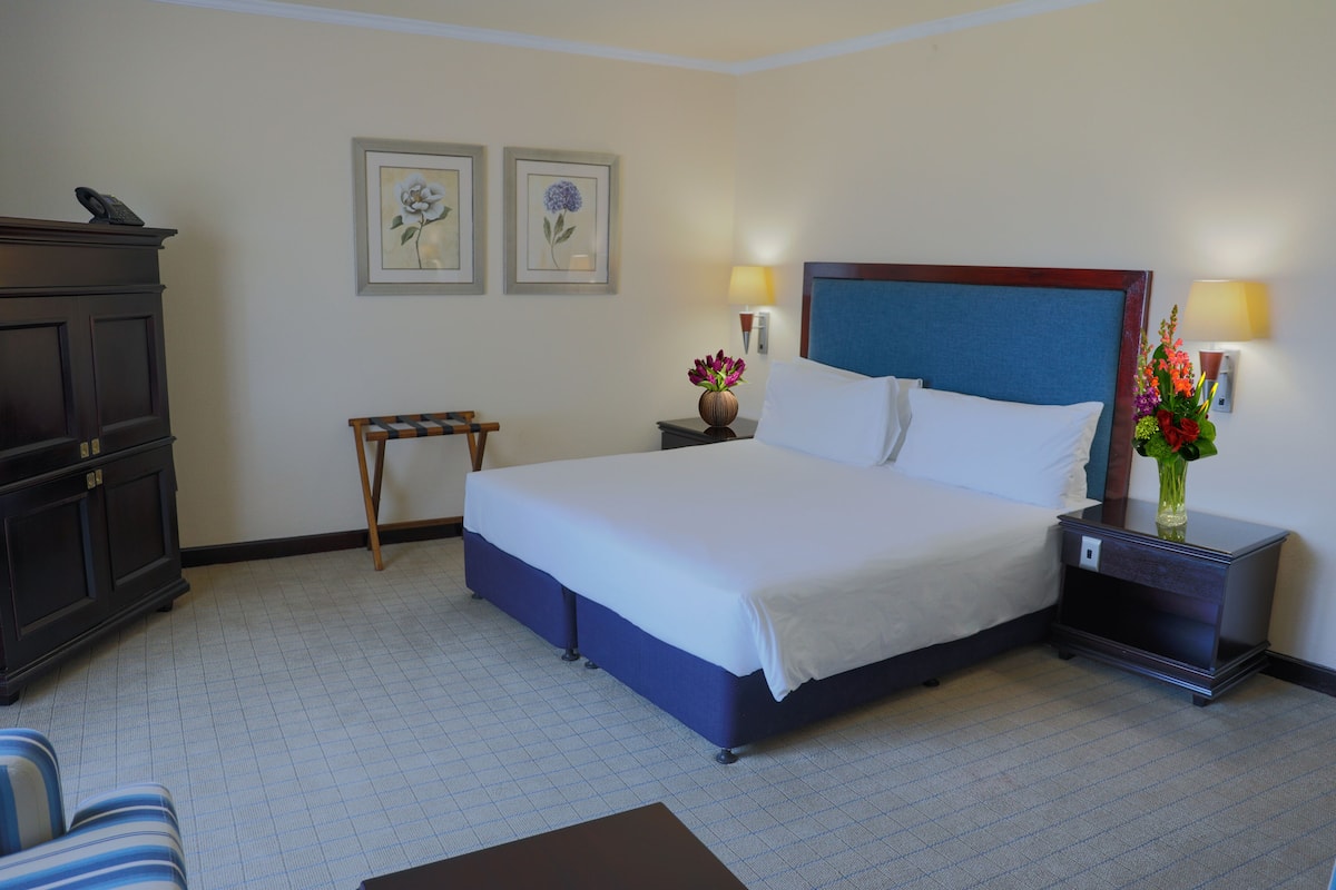 Een hotelkamer met een groot bed, een nachtkastje met lamp, een dressoir en twee bloemrijke foto’s in lijsten aan de muur.