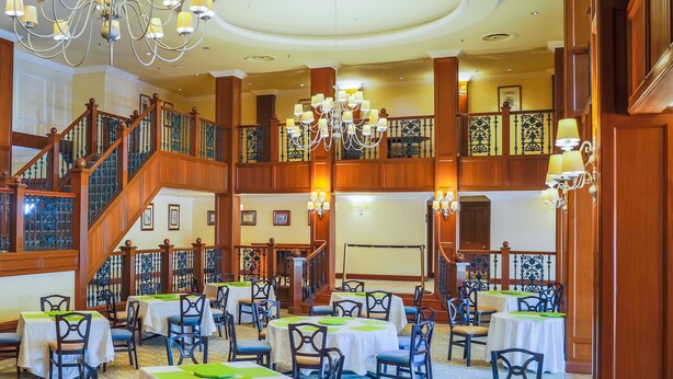 A spacious banquet hall with multiple round tables and chairs, a grand staircase, and ornate chandeliers. - The Carousel Hotel (Hammanskraal)