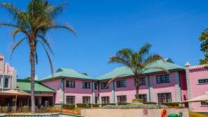 A resort with a swimming pool, palm trees, and pink buildings. - The Carousel Hotel (Hammanskraal)