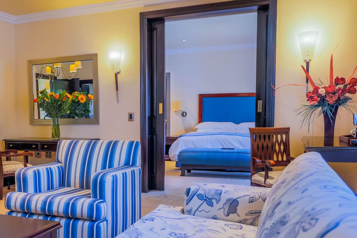 Hotelkamer met een bed, een gestreepte bank, een eettafel met stoelen, een kroonluchter en een grote spiegel.