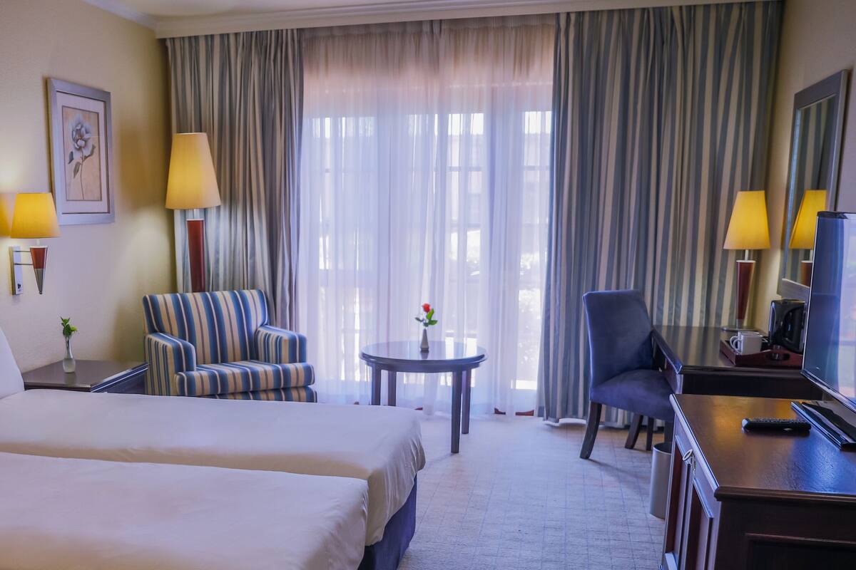 Een hotelkamer met twee bedden, een gestreept fauteuil, een klein rond tafeltje, een bureau met een computer en een lamp, en een groot raam met gordijnen.