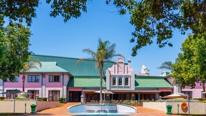 A large, colorful building with a green roof and multiple windows, surrounded by trees and a small pool. - The Carousel Hotel (Hammanskraal)