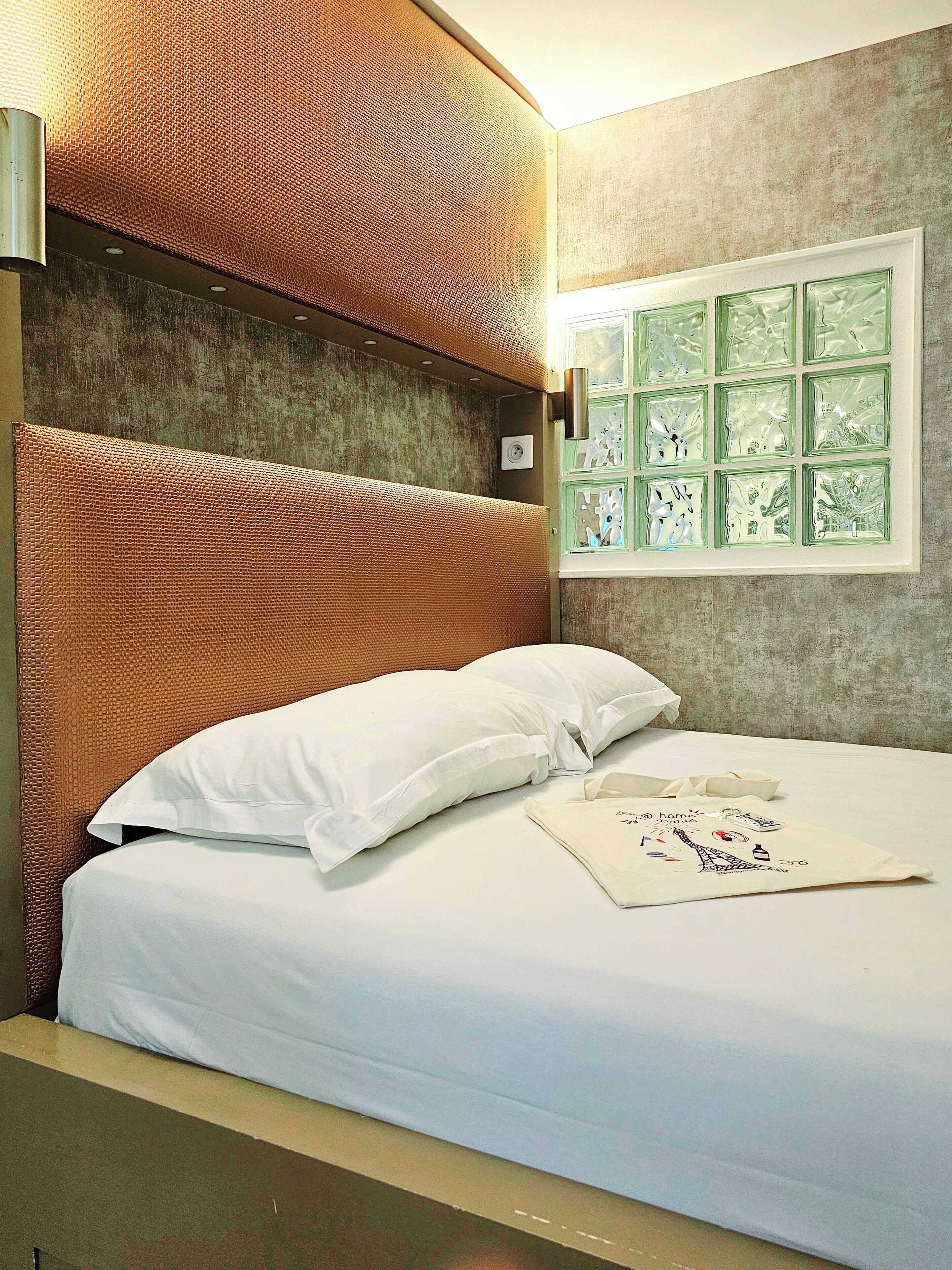 Cama bem arrumada com roupa de cama e travesseiros brancos, luminária fixada na parede e janela com painel de vidro decorativo.
