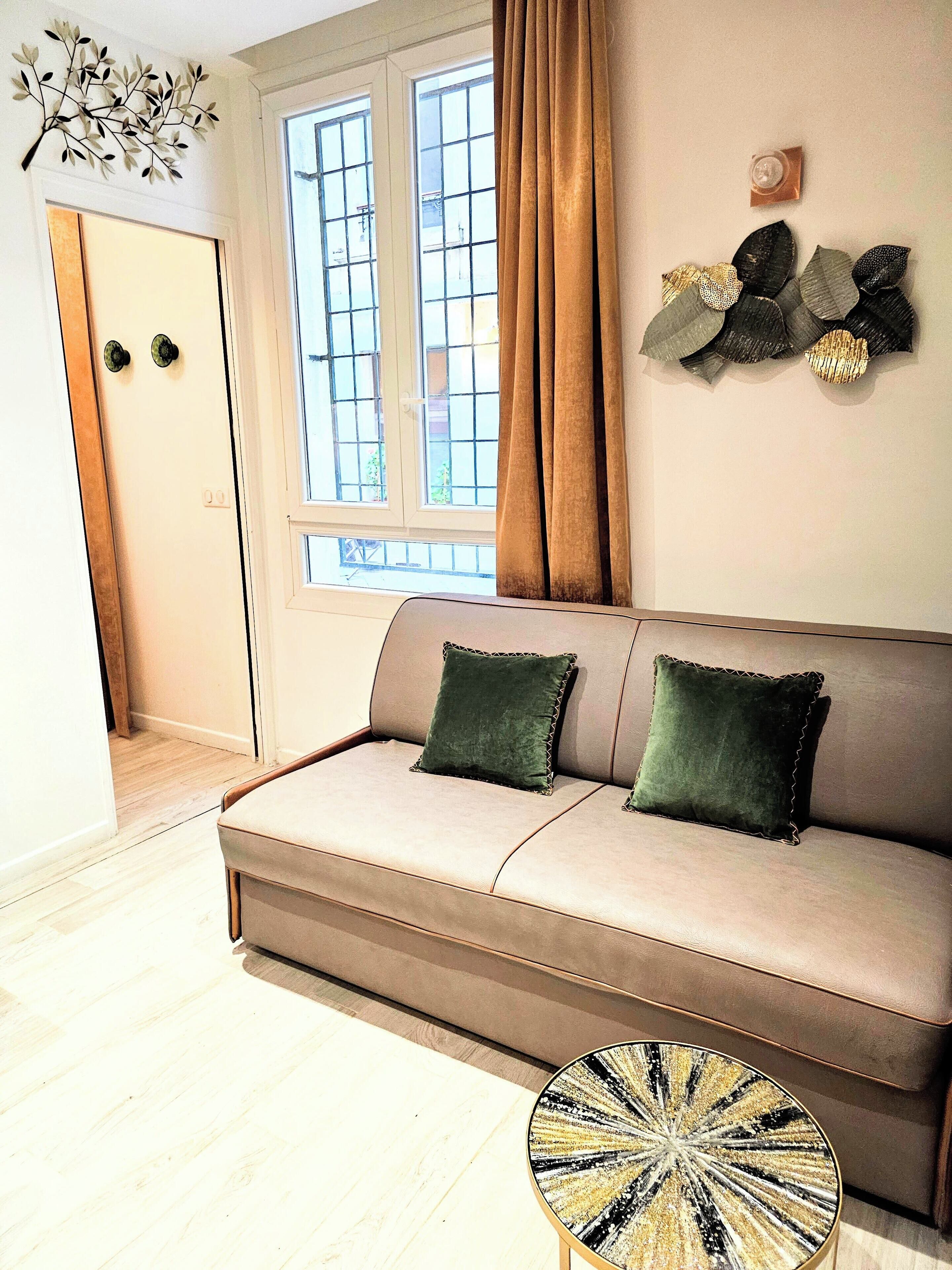 Una sala de estar con un sofá color beige, dos almohadas verdes, una mesa de centro redonda con una superficie decorativa, una ventana con cortinas y una obra de arte colgada en la pared.