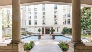 A courtyard with a central pool, surrounded by potted plants, and a multi-story building with balconies in the background. - Chic Designer Loft in Historic Mansion • Marais (Paris)
