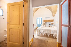Una habitación con puerta de madera, cama con cubrecama blanco, ventana con contraventanas azules y un arco de piedra.