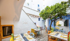 Un patio con mobiliario exterior, un árbol y edificios blancos con detalles azules.