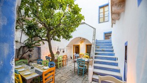 Un espace extérieur aménagé pour les repas, avec des chaises et des tables aux couleurs vives, un arbre et un escalier dont les marches sont bleues.