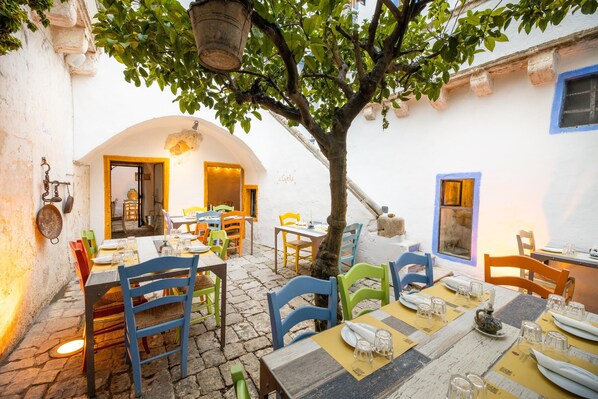 An outdoor dining area with tables and chairs, a tree, and a building with a white facade and blue trim. - Camera Onda Dorata - Racale (Racale)