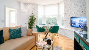 Ein modernes Wohnzimmer mit einem beigefarbenen Sofa, grünen Sesseln, einem Esstisch und einem Fernseher, der eine Stadtansicht zeigt.