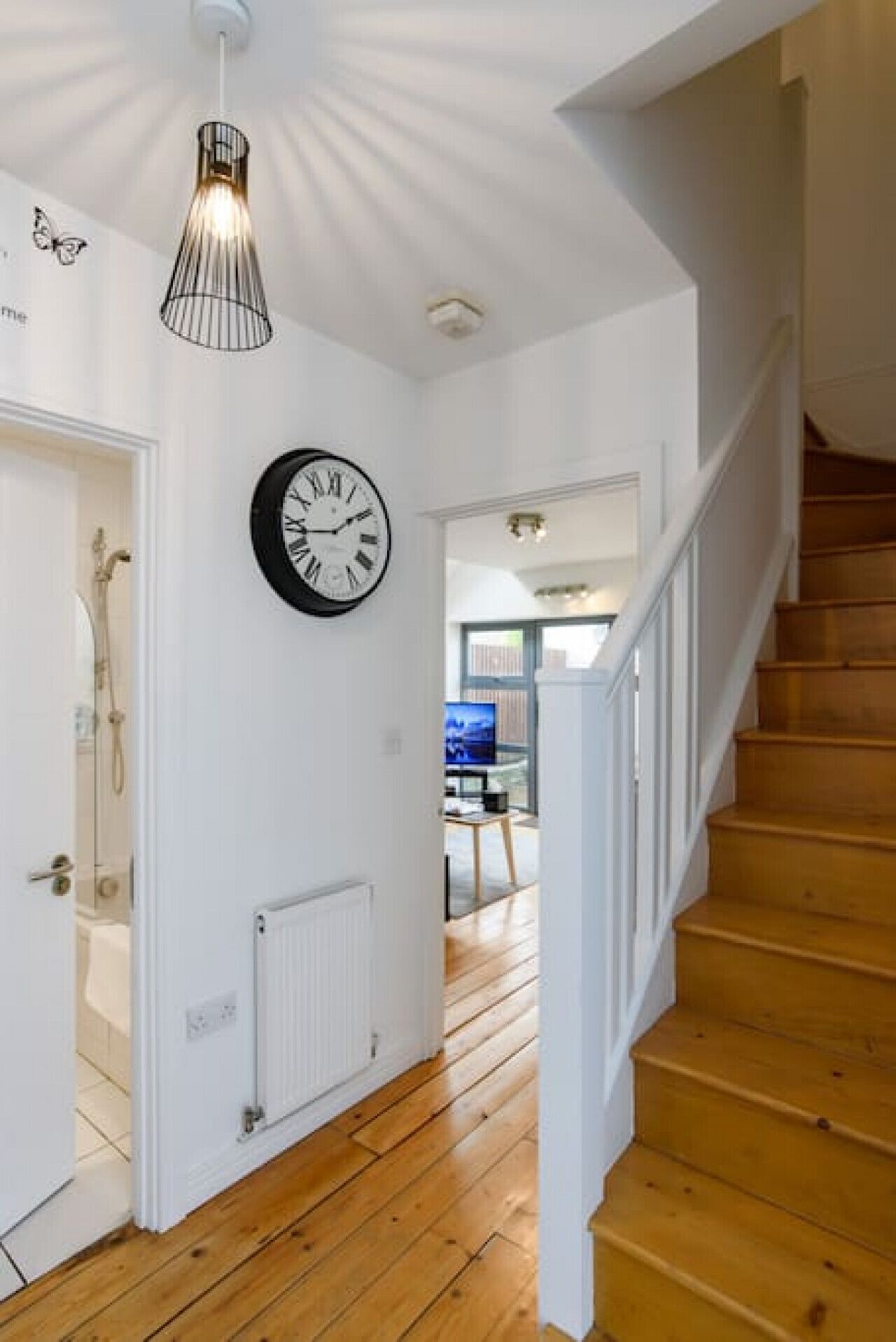 A staircase leading to a second floor with a wooden floor and white walls. A black clock is mounted on the wall, and a pendant light hangs above.
