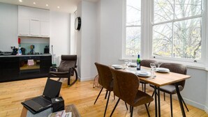 A modern kitchen with white cabinets, a dining area with a wooden table and chairs, and a large window with a view of trees. - Superb Modern 1 Bedroom Apt | Cocoon Dbl Sofa Bed (Cardiff)