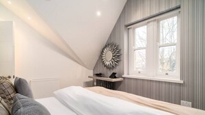 Een slaapkamer met een schuine kap, een bed, een nachtkastje met spiegel, en een raam met uitzicht op bomen.