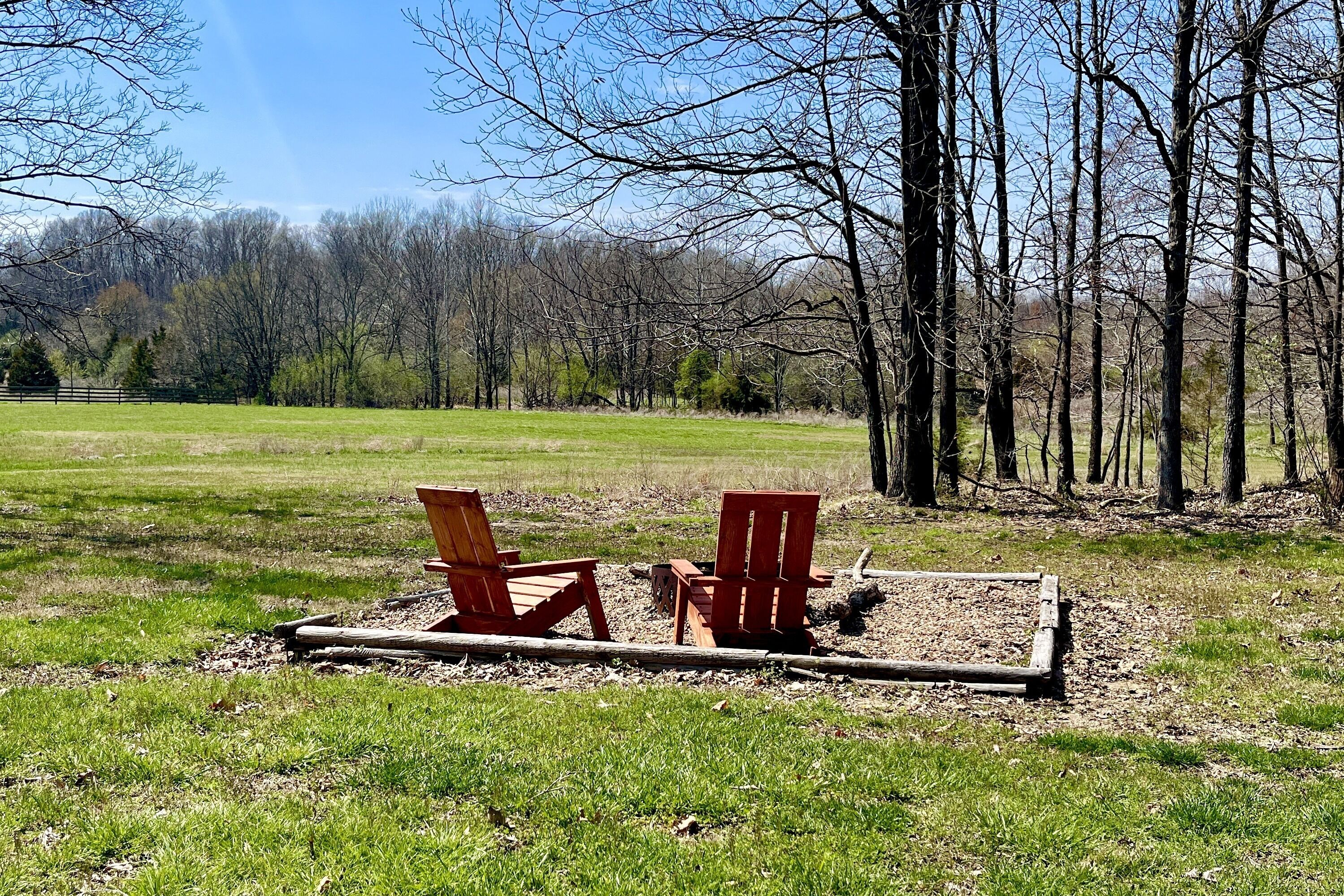 Two red Adirondack chairs in a grassy field with a wooden bench and a wooden fence.