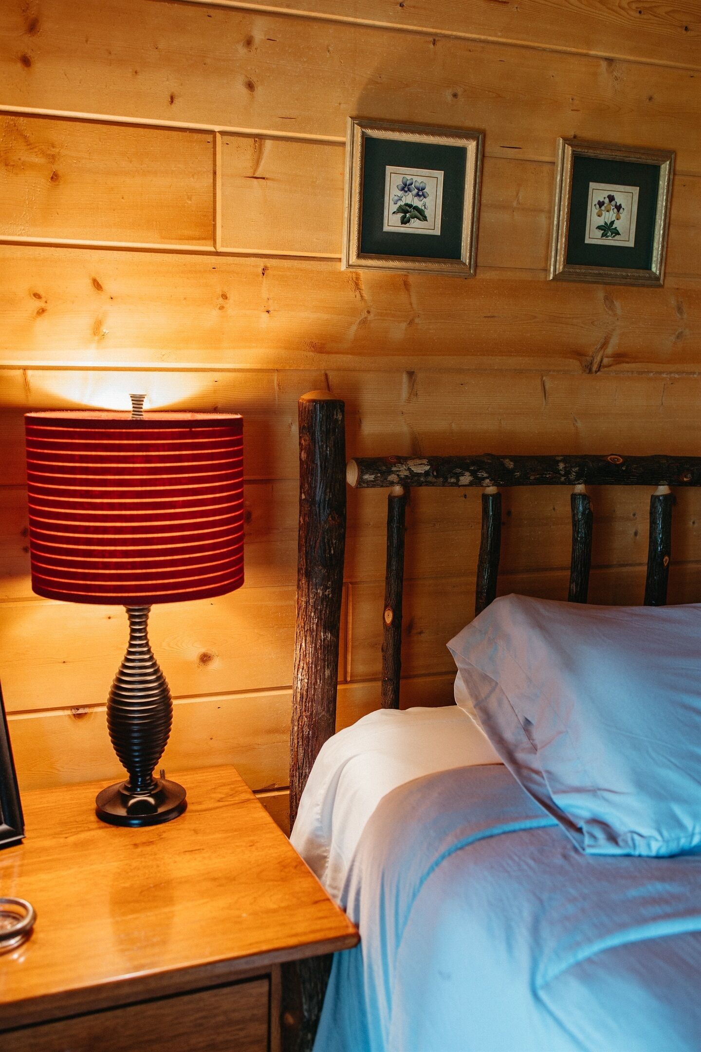 Una cama de madera con ropa blanca de cama, una lámpara rayada de rojo sobre una mesita de noche de madera, y dos cuadros con motivos florales en la pared.