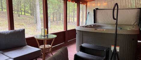 A covered outdoor seating area with a hot tub, wooden deck, and large windows overlooking a forest.