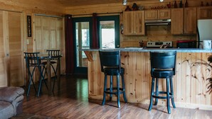 A wooden cabin interior with a kitchen area featuring a bar counter, stools, and wooden cabinets.