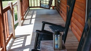 A wooden cabin with a porch, featuring two rocking chairs, a metal table, and a hanging lantern.