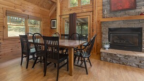 A wooden interior with a dining table and chairs, a fireplace, and large windows.