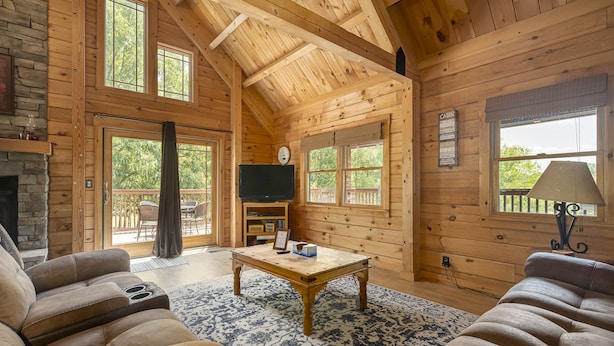 A log cabin interior with a fireplace, a television, a coffee table, and a large window.