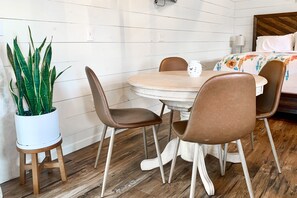 A small dining area with a round table and four chairs, a potted plant, and a bed with a floral bedspread in the background.