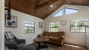 A cozy room with a brown leather sofa, a gray armchair, and a round coffee table. There are two windows with blinds, a ceiling fan, and a framed picture on the wall. - Upscale Cabin w/ Beach Near Lake of Egypt (Marion)