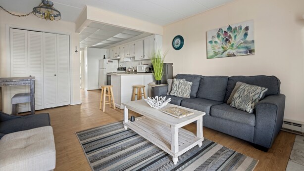 Ett vardagsrum med en grå soffa, ett vitt soffbord och en randig matta. I bakgrunden finns ett kök med vita skåp och en matplats.