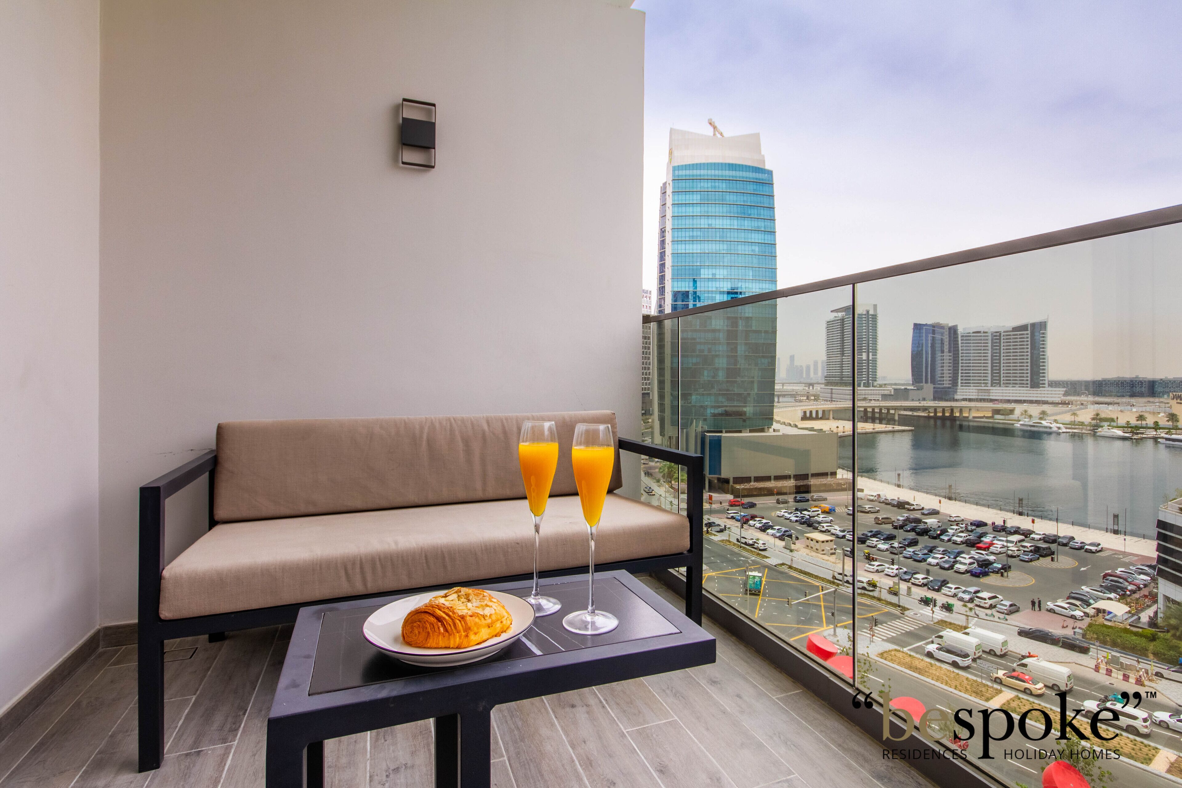 A balcony with a sofa, a table with a croissant and two glasses of orange juice, and a city view.