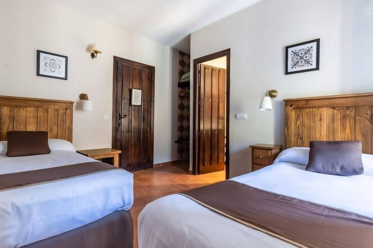A hotel room with two beds, wooden headboards, bedside tables, and wall-mounted lamps.