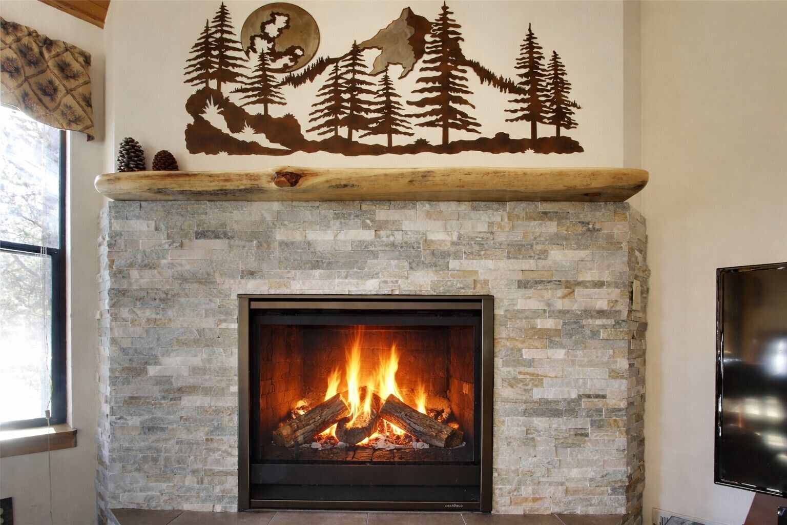 Une cheminée en pierre avec un feu de bois, un téléviseur fixé au mur et un décor mural sur le thème de la forêt.