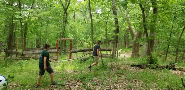 Deux personnes se trouvent dans une zone boisée, près d’une structure en bois et d’un pont suspendu.