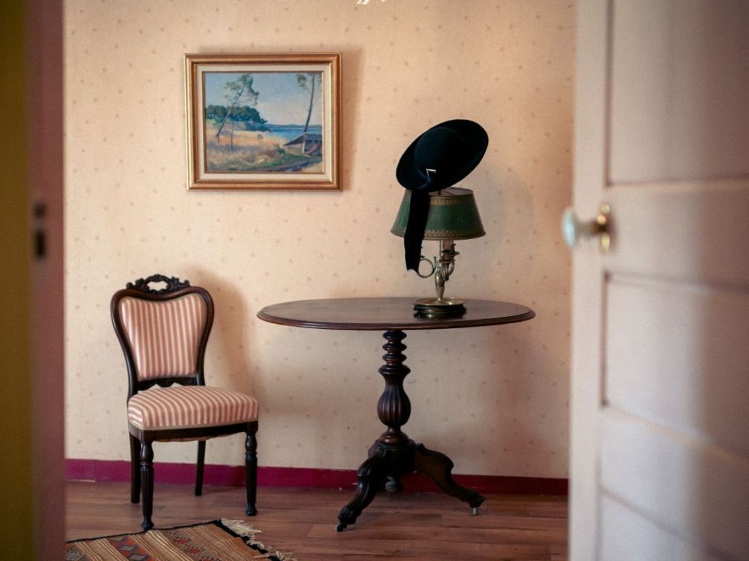 A room with a round table, a chair, a lamp, and a framed painting on the wall.