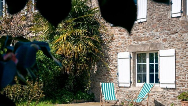 Un bâtiment en pierre avec un jardin, deux chaises de jardin à rayures et un palmier.