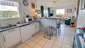 A modern kitchen with a built-in microwave, a sink, and a bar stool. There is a dining area with a table and chairs, and a view of the outdoors through a window. - Ker Mignonette - Trevou Treguignec (TREVOU TREGUIGNEC)