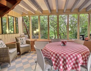 A cozy dining area with a checkered tablecloth, wicker chairs, and a view of lush greenery through large windows. - Ker Désirée - Plelo - Gîte (PLELO)