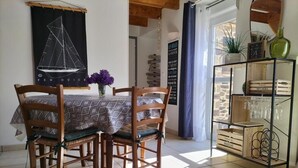 Ein Essbereich mit einem Tisch, bedeckt mit einer gemusterten Tischdecke, umgeben von Holzsesseln mit Kissen. An der Wand hängt ein Kunstwerk mit einem Segelboot, und ein Regal mit Gläsern und einer grünen Flasche ist sichtbar.