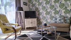 Ein Wohnzimmer mit einer Couch, einem gelben Sessel, einem Glastisch, einem Fernseher auf einer weißen Kommode und einer Wand mit Muster.