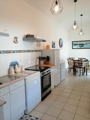Une cuisine moderne avec des meubles blancs, une cuisinière et un lave-vaisselle. On y trouve des luminaires suspendus et un coin repas avec une table et des chaises.