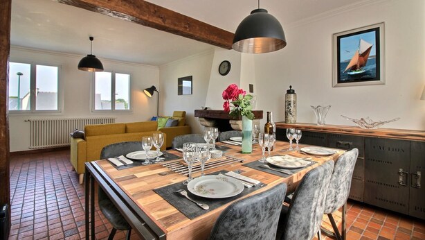 Un coin repas avec une table en bois dressée pour six personnes, entourée de chaises à revêtement gris. Un grand luminaire suspendu noir est accroché au plafond, et un tableau encadré représentant un voilier est accroché au mur.