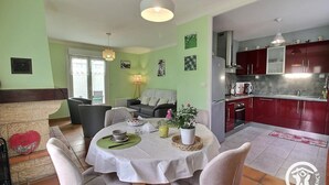 A dining area with a round table, chairs, and a small potted plant. A kitchen with red cabinets and a sink is visible in the background. - Le Jardin - Plestin LES Greves (PLESTIN LES GREVES)