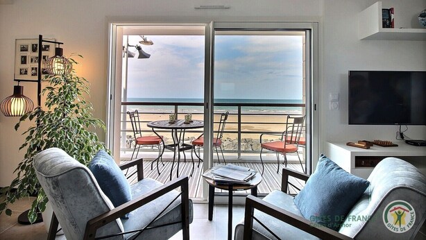 Una sala de estar moderna con un amplio ventanal con vistas a la playa, amueblada con un sofá, sillas y una mesita.