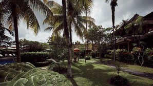 A tropical garden with palm trees, a pathway, and a building with a thatched roof. - Outpost Ubud Coliving Suites (Ubud)